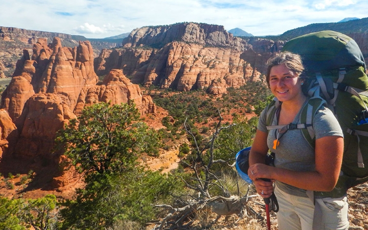 A person wearing a backpack smiles in front of a desert landscape with large red rock formations.